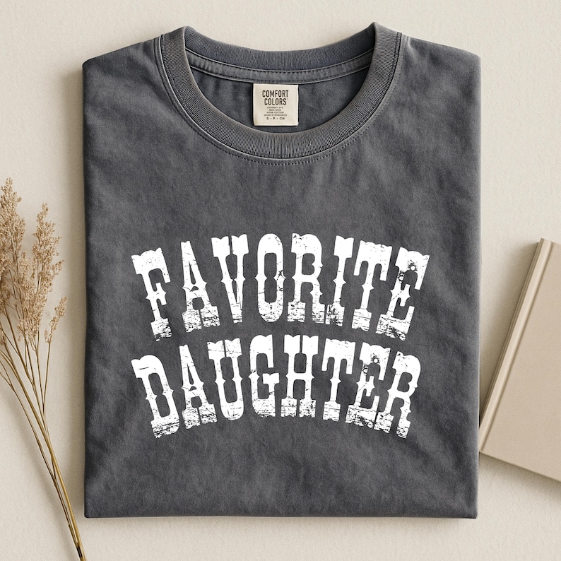 May include: A charcoal gray short-sleeve t-shirt with a crew neckline. The front features the white distressed text "FAVORITE DAUGHTER" in a bold, arched font. The interior neck label displays the brand name Comfort Colors and size S.