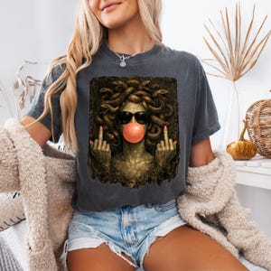 May include: A charcoal gray t-shirt featuring a graphic print of Medusa wearing black sunglasses and blowing a pink bubble. The figure has snakes for hair, is making a middle finger gesture with both hands, and is set against a dark background.