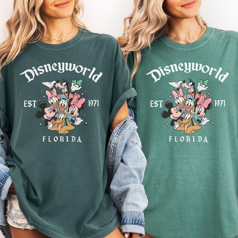 Puede incluir: Dos camisetas verdes con el texto "Disneyworld" y la frase "EST 1971 FLORIDA" en blanco. Las camisetas presentan un gr&aacute;fico de Mickey Mouse, Minnie Mouse, Donald Duck, Daisy Duck, Goofy y Pluto.