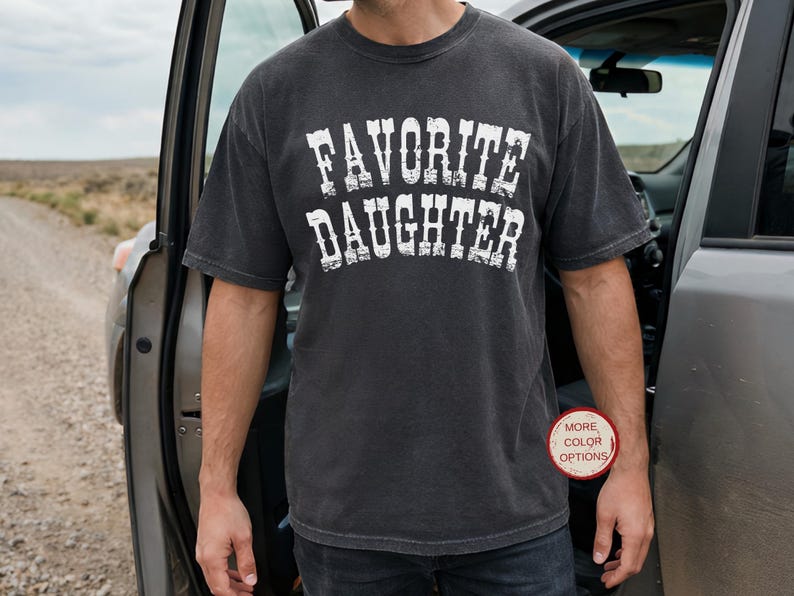 May include: A charcoal gray short-sleeve t-shirt with a crew neckline. The front features the text "FAVORITE DAUGHTER" printed in a distressed white font. A small circular badge in the lower right corner contains the text "MORE COLOR OPTIONS".