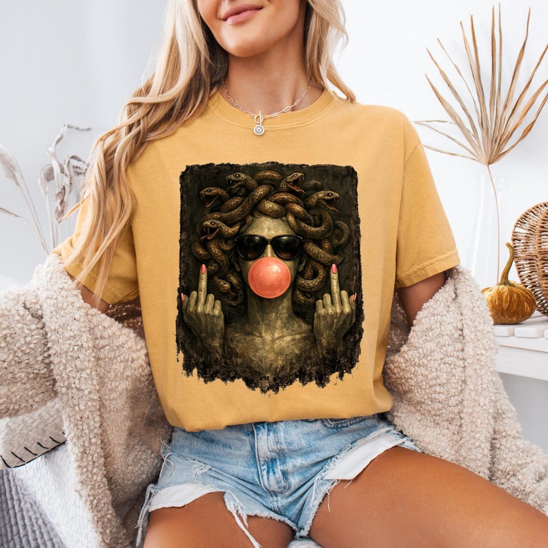 May include: A mustard yellow t-shirt featuring a graphic print of a figure with snakes for hair, wearing black sunglasses and blowing a pink bubblegum bubble. The figure has green skin and is shown making a middle finger gesture with both hands.