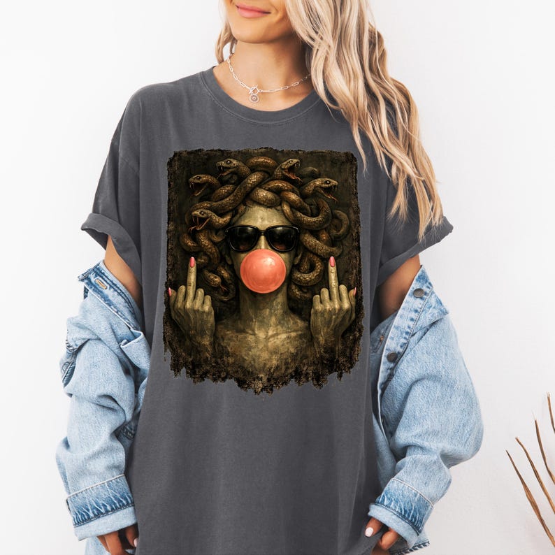 May include: A charcoal gray t-shirt featuring a graphic print of a Medusa figure with snakes for hair, wearing black sunglasses and blowing a pink bubblegum bubble. The figure is shown with both middle fingers raised in a defiant gesture.