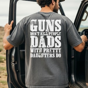 May include: A charcoal gray short-sleeve t-shirt featuring white distressed-style text on the back that reads: GUNS DON'T KILL PEOPLE DADS WITH PRETTY DAUGHTERS DO. The shirt has a crew neckline and a relaxed fit, showcasing a vintage-inspired wash.