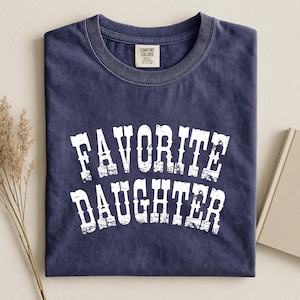 May include: A navy blue short-sleeved t-shirt with a crew neckline. The shirt features the white distressed text "FAVORITE DAUGHTER" printed in a bold, arched font across the chest. A Comfort Colors brand label is visible at the inner neck.