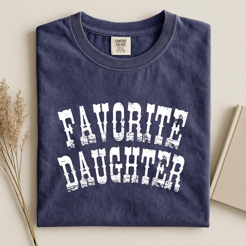 May include: A navy blue short-sleeved t-shirt with a crew neckline. The shirt features the white distressed text "FAVORITE DAUGHTER" printed in a bold, arched font across the chest. A Comfort Colors brand label is visible at the inner neck.