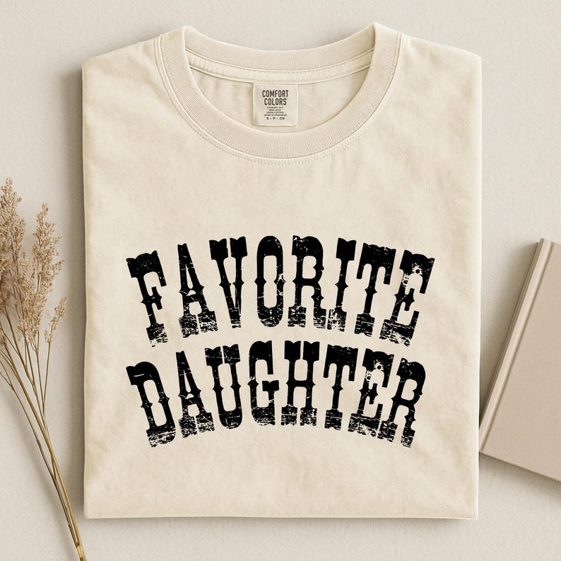 May include: A cream-colored short-sleeve t-shirt with a crew neckline. The front features the text "FAVORITE DAUGHTER" printed in a black, distressed, arched western-style font. A Comfort Colors brand label is visible at the inner neck area.