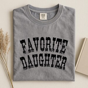 May include: A grey short-sleeved crew neck t-shirt featuring the black distressed text "FAVORITE DAUGHTER" printed in a curved, western-style font across the chest. The shirt includes a Comfort Colors brand label at the inner neckline.