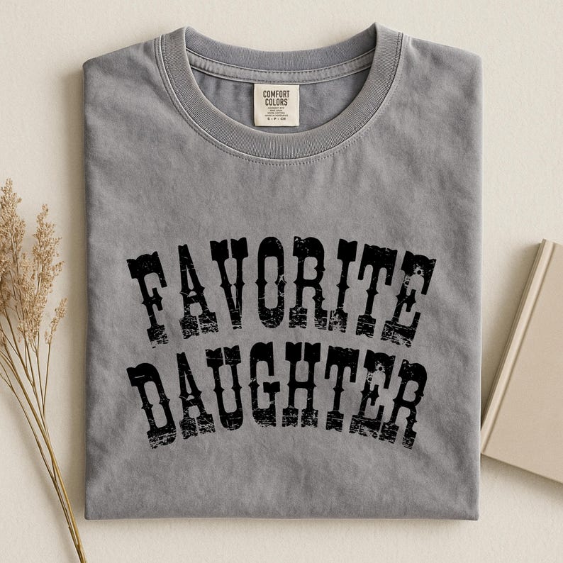 May include: A grey short-sleeved crew neck t-shirt featuring the black distressed text "FAVORITE DAUGHTER" printed in a curved, western-style font across the chest. The shirt includes a Comfort Colors brand label at the inner neckline.