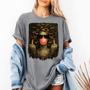 May include: A grey t-shirt featuring a graphic print of a Medusa figure with snakes for hair, wearing black sunglasses and blowing a pink bubblegum bubble. The figure is shown with both middle fingers raised in a gesture against a dark background.