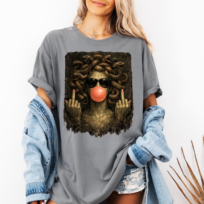 May include: A grey t-shirt featuring a graphic print of a Medusa figure with snakes for hair, wearing black sunglasses and blowing a pink bubblegum bubble. The figure is shown with both middle fingers raised in a gesture against a dark background.