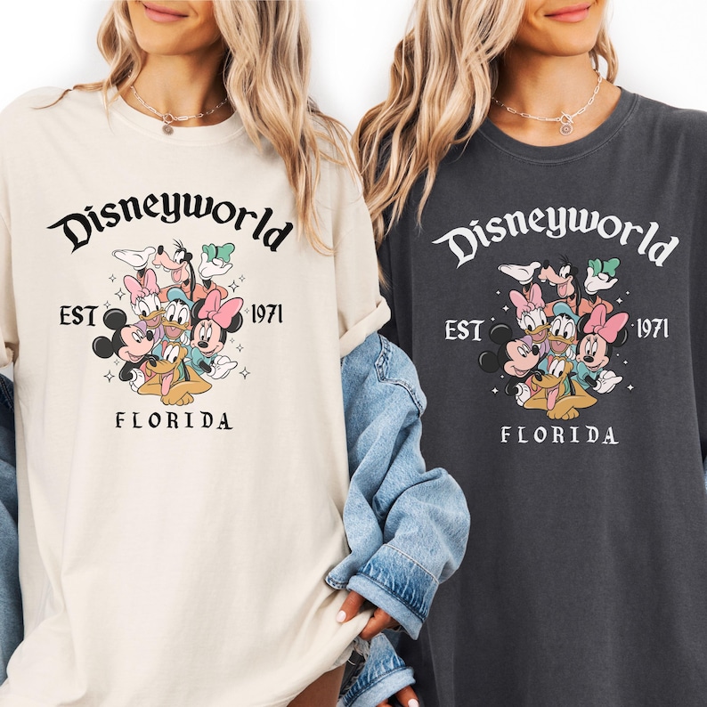 Puede incluir: Dos camisetas, una crema y otra antracita, con el texto "Disneyworld" sobre un gr&aacute;fico de personajes de Disney. Debajo, las palabras "EST 1971 FLORIDA". Las camisetas tienen las mangas enrolladas.