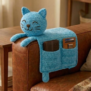 Puede incluir: Un organizador de reposabrazos de sofá con forma de gato tejido en crochet azul claro. El gato tiene una cara sonriente y dos orejas pequeñas. El organizador tiene dos bolsillos y está colocado sobre un reposabrazos de sofá de cuero marrón. Se ven carteras.