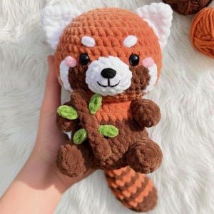 May include: A handmade crochet red panda plush toy. The plush features a round head with white ears, a white muzzle, and black eyes. It has a brown body with orange accents and holds a brown branch with green leaves. The tail is striped.