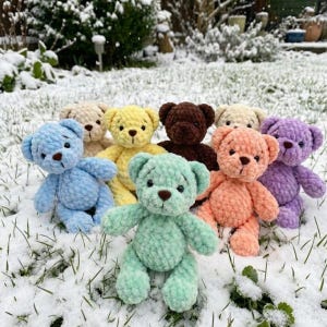 May include: A group of colourful, handmade crochet teddy bears. The bears are in shades of blue, yellow, brown, peach, and purple, with brown button eyes. They are arranged on a snow-covered surface, highlighting their soft, textured appearance.