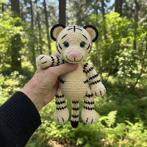 May include: A crocheted tiger plush toy with cream-colored yarn and black stripes. The tiger has a pink nose, blue eyes, and black accents on the ears and paws. The handmade stuffed animal is held in front of a blurred forest background.