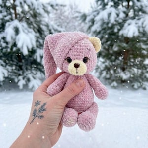 Op de afbeelding: Een handgemaakte roze teddybeer met een bijpassende nachtmuts. De beer heeft een lichtbeige snuit en oren, met donkerbruine ogen en een gestikte neus. Het pluche speelgoed wordt vastgehouden tegen een wazige, besneeuwde achtergrond van naaldbomen.