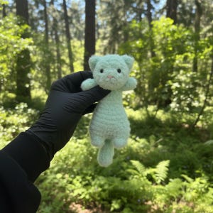 May include: A small, mint green crocheted cat doll with a fluffy texture. The cat has small, round ears, a pink nose, and black eyes. The doll is held against a backdrop of a lush green forest.