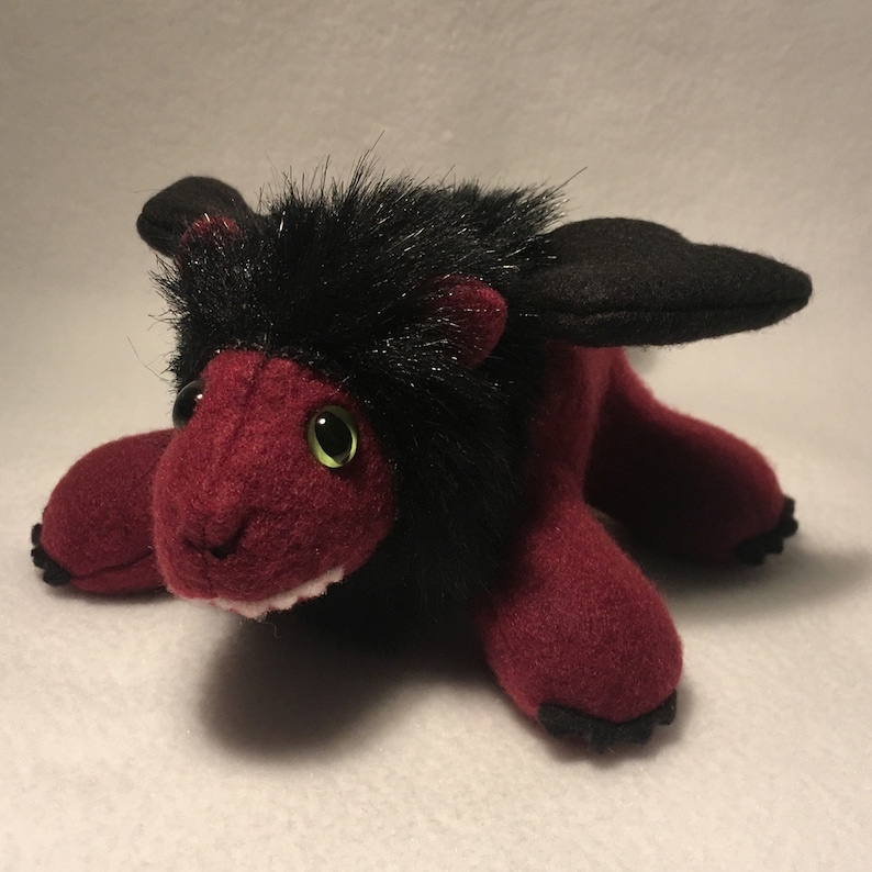 May include: A plush toy in the shape of a red and black winged lion with green eyes. The toy has a toothy grin and black fur on its head.