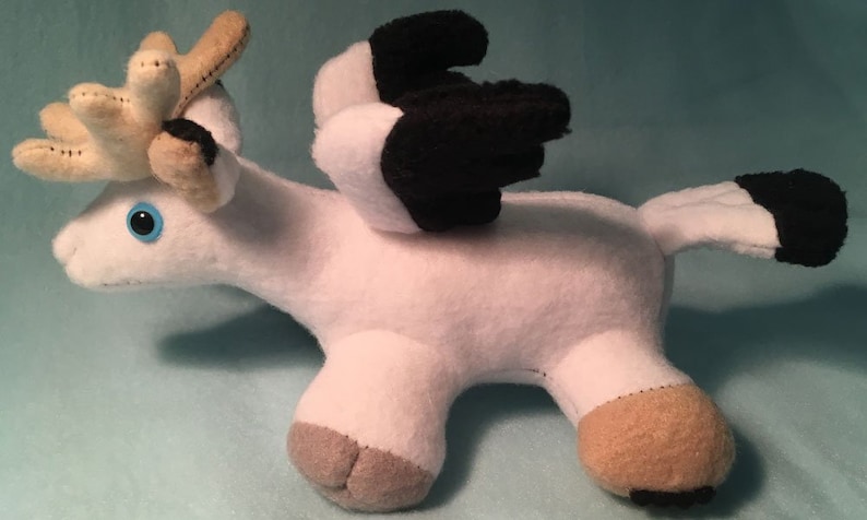 K&ouml;nnte beinhalten: Wei&szlig;es Pl&uuml;schtier mit einem Hirschkopf, schwarzen Fl&uuml;geln und einem braunen Schwanz. Das Spielzeug hat blaue Augen und eine braune Nase.