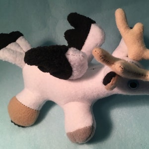 K&ouml;nnte beinhalten: Wei&szlig;es Pl&uuml;schtier mit schwarzen Fl&uuml;geln, braunen Geweih und blauen Augen. Das Spielzeug befindet sich in einer Flugpose.