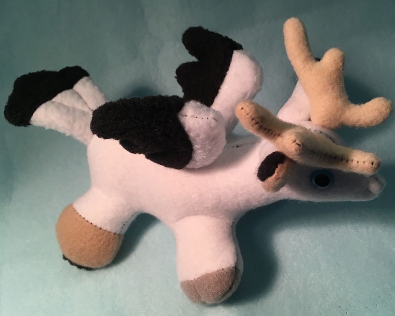 K&ouml;nnte beinhalten: Wei&szlig;es Pl&uuml;schtier mit schwarzen Fl&uuml;geln, braunen Geweih und blauen Augen. Das Spielzeug befindet sich in einer Flugpose.