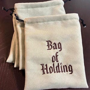 May include: A collection of light green suede-like drawstring bags. Each bag features the embroidered words "Bag of Holding" in a dark brown, gothic font. The drawstrings are black.