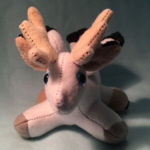 K&ouml;nnte beinhalten: Ein wei&szlig;es Pl&uuml;schtier mit braunen Geweihen und blauen Augen. Das Spielzeug liegt auf dem Bauch mit ausgestreckten Beinen.