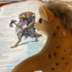 May include: A brown plush hyena toy with dark brown spots and red eyes. It has a gray mane and is positioned in front of an open book. The book features text and an illustration of a fantasy creature.