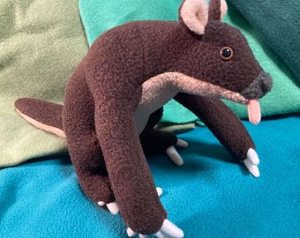 May include: A brown plush toy animal with a long snout and a pink tongue sticking out. The toy has a brown body with a lighter brown snout and ears. The toy is sitting on a blue surface.
