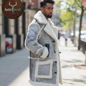 May include: A light gray shearling coat with a white shearling lining and trim. The coat features a wide collar, large pockets, and black accents. The coat is buttoned up and worn by a person standing on a city sidewalk.