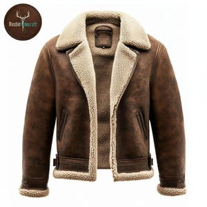 Peut inclure: Veste en cuir marron avec une doublure et un col en peau de mouton crème. La veste est dotée d'une fermeture éclair, de deux poches avant et de poignets réglables. Le logo "Rusticdecraft" est visible en haut à gauche.