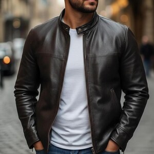 K&ouml;nnte beinhalten: Eine dunkelbraune Lederjacke mit Stehkragen, einem zentralen Metallrei&szlig;verschluss und langen &Auml;rmeln. Die Jacke wird &uuml;ber einem schlichten wei&szlig;en T-Shirt und einer blauen Jeans getragen. Das Leder hat eine glatte Oberfl&auml;che mit sichtbaren N&auml;hten.