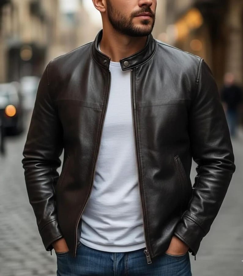 K&ouml;nnte beinhalten: Eine dunkelbraune Lederjacke mit Stehkragen, einem zentralen Metallrei&szlig;verschluss und langen &Auml;rmeln. Die Jacke wird &uuml;ber einem schlichten wei&szlig;en T-Shirt und einer blauen Jeans getragen. Das Leder hat eine glatte Oberfl&auml;che mit sichtbaren N&auml;hten.