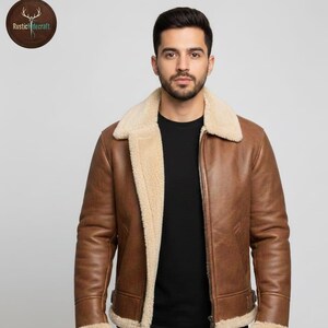 Könnte beinhalten: Braune Lederjacke mit cremefarbenem Shearling-Futter und -Kragen. Die Jacke hat einen Reißverschluss und Schnallendetails an der Taille. Das Rustic Hidecraft-Logo ist oben links sichtbar.