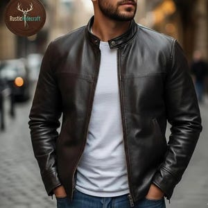 K&ouml;nnte beinhalten: Eine dunkelbraune Lederjacke mit Stehkragen, Frontrei&szlig;verschluss und einem Druckknopf am Hals. Die Jacke wird &uuml;ber einem schlichten wei&szlig;en T-Shirt und einer blauen Jeans getragen. Ein kreisf&ouml;rmiges Logo mit dem Text RusticHidecraft ist sichtbar.