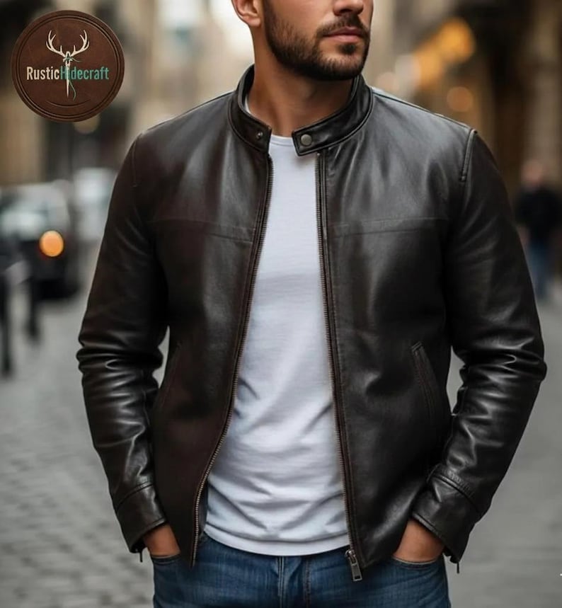 K&ouml;nnte beinhalten: Eine dunkelbraune Lederjacke mit Stehkragen, Frontrei&szlig;verschluss und einem Druckknopf am Hals. Die Jacke wird &uuml;ber einem schlichten wei&szlig;en T-Shirt und einer blauen Jeans getragen. Ein kreisf&ouml;rmiges Logo mit dem Text RusticHidecraft ist sichtbar.