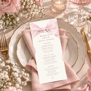 May include: A tall, rectangular menu card for a bridal shower featuring a soft pink ribbon illustration at the top. The text reads: MENU, OLIVIA'S, A Toast to Love, BRIDAL SHOWER, STARTERS, MAIN COURSE, and DESSERT, followed by various food items.