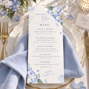 May include: A rectangular paper menu featuring blue and white hydrangea floral illustrations at the top and bottom. The text reads Something Blue Menu, listing courses including Burrata, Lemon Herb Risotto, Herb Crusted Salmon, Filet Mignon, and Vanilla Bean Panna Cotta.