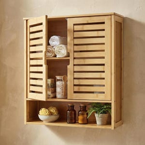 May include: A light brown bamboo wall-mounted cabinet with slatted doors, open to reveal rolled towels, jars, and soaps. The lower shelf holds a bowl, two brown bottles, and a potted plant. The cabinet is designed for bathroom storage.
