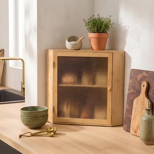 May include: A corner cabinet made of light-toned bamboo wood featuring a single door with a fluted glass panel. The cabinet has a rectangular frame with angled sides, a small round wooden knob, and two internal shelves visible through the glass.