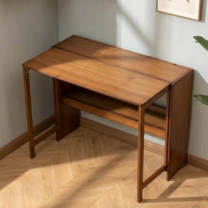 May include: A rectangular wooden desk with a warm brown finish, featuring a smooth tabletop surface, a lower horizontal storage shelf, and a hinged side panel that allows for folding, supported by four straight legs and a solid side panel base.
