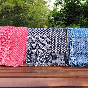May include: Three patterned textiles in red, navy, and blue are folded on a wooden bench. The red textile has a floral design, the navy one has a white leaf pattern, and the blue one has a circular design. The textiles are outdoors.