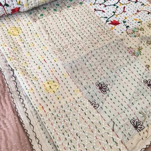 May include: A white cotton quilt featuring a patchwork design with intricate kantha stitching. The fabric displays various embroidered floral patterns in red, yellow, and green, alongside sections of white lace and decorative geometric embroidery.