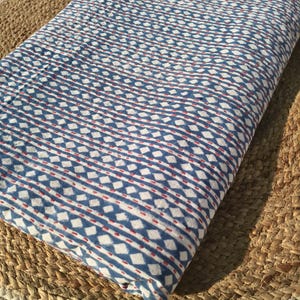 May include: Folded fabric with a blue and white geometric pattern, accented with red dashes. The fabric is likely cotton or a similar material, and the design features diamond and line motifs. The fabric is resting on a woven, natural-toned surface.