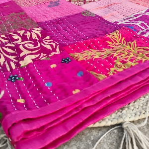 May include: A patchwork quilt in shades of pink and magenta. The quilt is made of various fabric squares with different patterns and textures, including floral and geometric designs. The edges are neatly finished with a magenta border. The quilt is folded, revealing its layered construction.