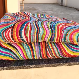 May include: Two folded, colorful quilts with a vibrant, swirled pattern of vertical stripes in shades of red, orange, yellow, green, blue, and purple. The quilts are stacked on top of each other, showcasing the intricate design and stitching.