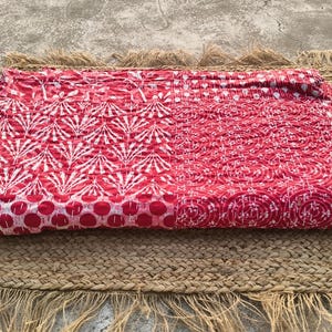 Peut inclure: Un textile plié à motifs rouges et blancs repose sur un tapis en jute tissé avec des bords frangés. Le textile présente un motif répétitif de motifs floraux blancs et circulaires sur fond rouge. Le tapis est de couleur beige naturel.