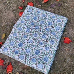May include: A rectangular piece of white fabric featuring a repeating pattern of blue floral clusters and green leafy vines. The design consists of circular arrangements of small blue dots surrounded by thin, winding brown stems and green leaves.