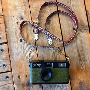 May include: A black and olive green film camera with a textured finish and the text "Camp Snap" on the front. The camera is attached to a long, thin fabric neck strap featuring a repeating geometric pattern in shades of red, white, and black.