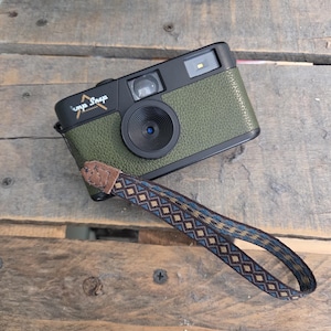 Peut inclure: Un appareil photo numérique Camp Snap avec un boîtier texturé vert olive et des finitions noires. L'appareil comprend un objectif central, un viseur et un flash. Il est muni d'une dragonne tissée avec une patte en cuir marron et un motif losange bleu, marron et doré.